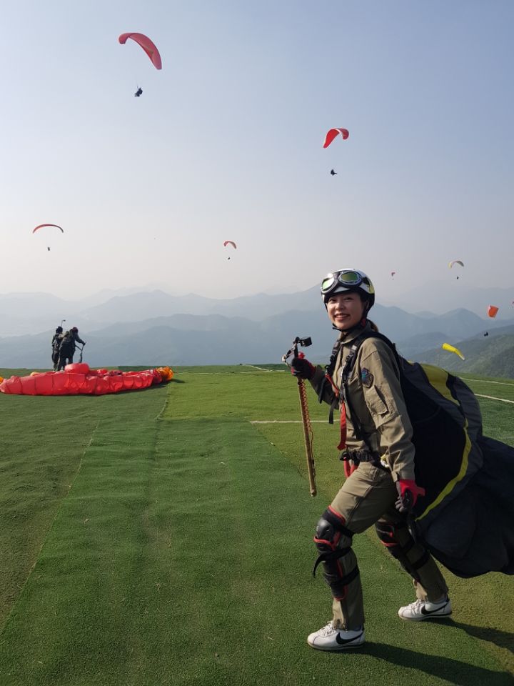 Rosie Lee in paragliding gear on a mountain launch field, with gliders in the sky behind her