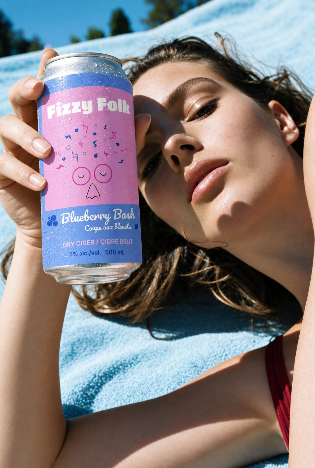 AI-generated scene: a woman lying on a pale blue towel in the sun holds up a chilled Fizzy Folk Blueberry Bash can