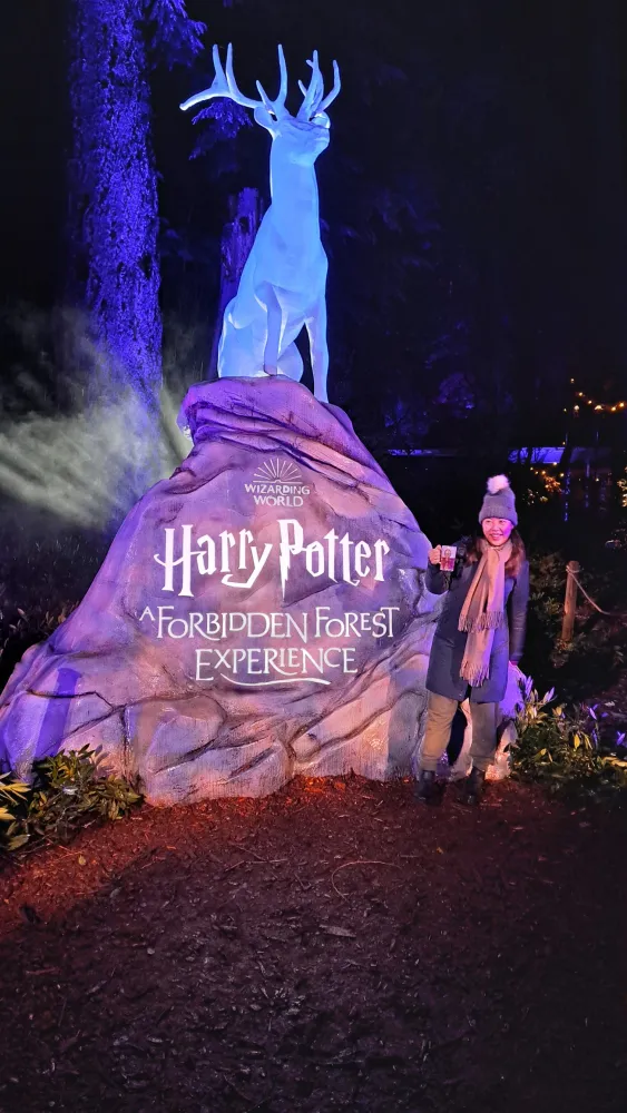 Rosie holding a mug beside the glowing stag statue at Harry Potter: A Forbidden Forest Experience, at night