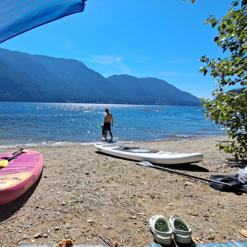 Rosie’s pink paddleboard and a white one on a pebble beach by a mountain lake, under a beach umbrella