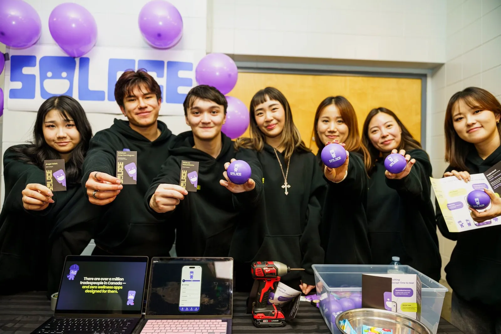 The seven-person Solace team at their showcase booth, holding up business cards, branded stress balls and the brochure