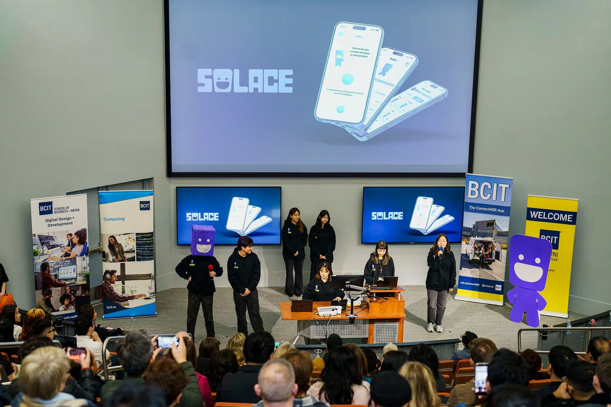 The Solace team pitching on stage at the BCIT Final Product Showcase, with the mascot costume, a life-size mascot cutout and the app on screen