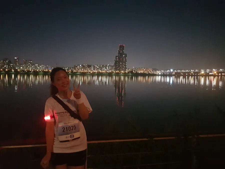 Rosie in a Hangang Night Walk T-shirt and race bib, making a peace sign by the Han River at night with the city lit up behind her