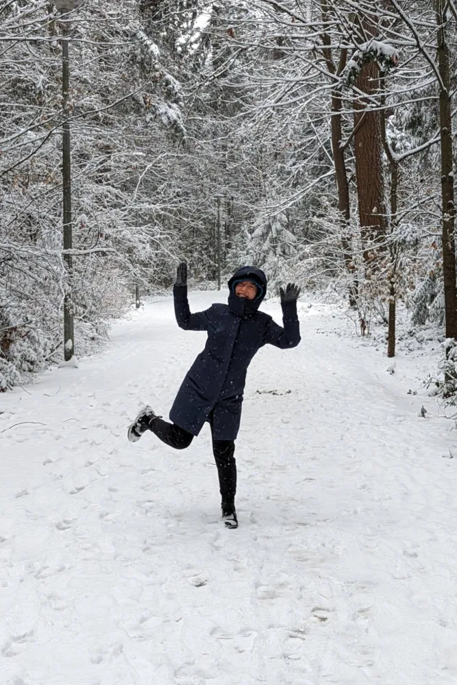 Rosie in a winter coat kicking up one leg and waving on a snowy forest path